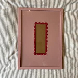 May include: A rectangular pink picture frame with a red scalloped border around the photo opening. The frame is set against a striped fabric background. The photo opening is a light brown color.