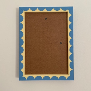 Könnte beinhalten: Ein rechteckiger Bilderrahmen mit einem blauen, wellenförmigen Rand und einem gelben Innenrand. Der Rahmen hat eine braune Rückseite und zwei kleine Löcher zum Aufhängen. Der Rahmen ist an einer weißen Wand befestigt.