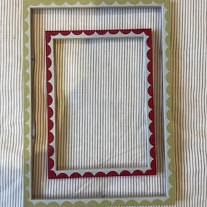 Può includere: Due cornici decorative. La cornice più grande è verde chiaro con un bordo smerlato. La cornice più piccola è rossa con un bordo smerlato. Entrambe le cornici sono rettangolari e poggiano su un tessuto a righe.