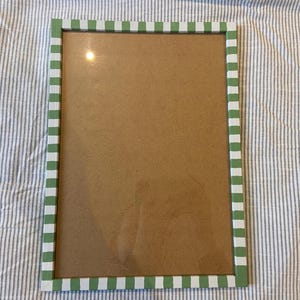 May include: Rectangular picture frame with a green and white checkered border. The frame has a clear glass front and a brown backing. The frame is resting on a striped fabric.