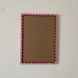 May include: A rectangular picture frame featuring a light pink outer border and a dark red inner border with a scalloped edge design. The center of the frame is filled with a flat, brown, solid-colored backing material, creating a simple display.