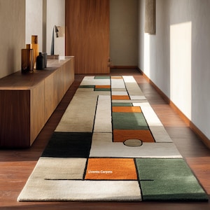 May include: A long rectangular runner rug featuring a geometric pattern of cream, dark green, black, and orange blocks outlined in thin black lines. The text Livento Carpets is printed in white on one of the orange rectangular sections of the rug.