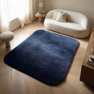 May include: A navy blue rectangular rug with rounded corners sits on a hardwood floor. The rug is the focal point of a living room setting, with a white curved sofa and a small side table visible in the background. The rug's texture appears soft and plush.