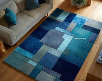 Puede incluir: Una alfombra rectangular con un patrón geométrico de cuadrados y rectángulos superpuestos en varios tonos de azul, verde azulado y gris. La alfombra muestra el texto Livento Carpets en letras blancas sobre su superficie de pelo texturizado.