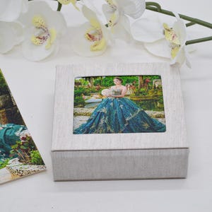 May include: A square, light beige fabric-covered keepsake box featuring a centered, inset photograph of a person wearing a voluminous, dark teal ball gown with gold embroidery, standing in a lush garden setting with a pond and greenery.