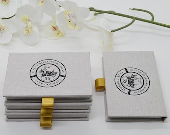 May include: A stack of light gray linen-textured boxes with gold ribbon pulls. Each box features a circular emblem with the text "City of Dearborn Michigan" and the number "35". White orchid flowers are in the background.