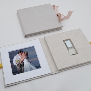 Peut inclure: Un album photo de mariage recouvert de lin gris clair avec une clé USB assortie. L'album est ouvert, révélant une photo de mariage encadrée et une clé USB. Un ruban rose est noué autour de l'album. Un souvenir pour les photos de mariage.