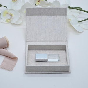 May include: An open, rectangular, light gray linen box with a hinged lid. Inside, two small, rectangular objects sit side-by-side. One is silver, the other is clear. White orchids and pink ribbon are in the background.