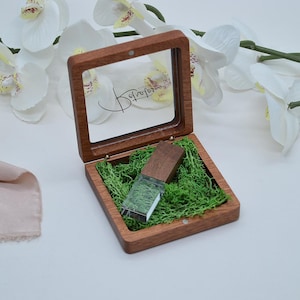 May include: A square wooden box with a hinged glass lid featuring the script text Retratos. Inside, a rectangular wooden USB drive rests on a bed of bright green preserved moss. The box has a natural wood grain finish and a magnetic closure.