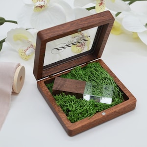 May include: Open wooden box with a clear lid, containing a wooden USB drive and a clear USB drive. The box is lined with green moss. The lid has a logo. White orchids are in the background.