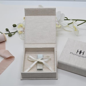 May include: Open, rectangular, linen-textured box with a silver object tied with a white ribbon. The box is open, revealing the contents. A smaller box with the text "HH Photography" is to the right.