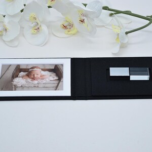 Peut inclure: Un coffret de présentation rectangulaire noir avec une photographie à bordure blanche d'un nouveau-né sur la gauche et une clé USB argentée insérée dans une mousse noire sur la partie droite de l'intérieur ouvert.