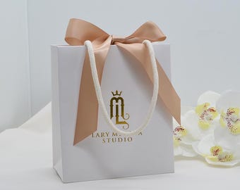May include: A white gift bag with a peach-colored satin bow and rope handles. The bag features gold lettering and a logo. A cluster of white orchids with yellow centers is in the background.