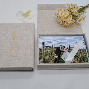 Op de afbeelding: Een linnen fotoalbum met de gouden tekst "HOLLY FLORENCE". Het album is open en toont een trouwfoto en een wit lint. Een klein boeket bloemen en een witte bloem zijn ook aanwezig.