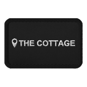 May include: Black rectangular patch with white embroidered text and a location pin graphic. The text reads "THE COTTAGE". The patch has a stitched border.