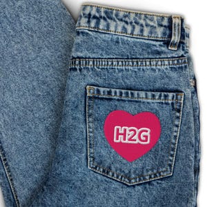 May include: Close-up of light blue denim jeans. The back pocket features a pink heart-shaped patch with the white embroidered letters "H2G". The jeans have a classic five-pocket design and a faded wash.