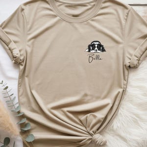 Könnte beinhalten: Beiges T-Shirt mit einer schwarz-weißen Hundezeichnung und dem Namen "Bella" darunter. Die Ärmel sind hochgekrempelt und das T-Shirt ist am Saum geknotet. Der Hintergrund ist eine flauschige, weiße Oberfläche.
