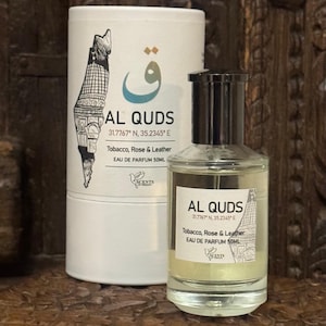 May include: A perfume bottle and cylindrical box with the text "AL QUDS" and "Tobacco, Rose & Leather" are displayed. The bottle is clear with a black cap. The box and bottle have a graphic of a building. The perfume is 50ml.