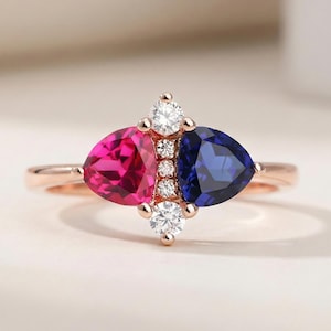 Op de afbeelding: Een roségouden ring met een opvallend ontwerp. Het heeft een fuchsia roze en een diepblauwe, driehoekig geslepen edelsteen, geaccentueerd door kleine, sprankelende witte stenen. De ring heeft een delicate, gepolijste roségouden band.