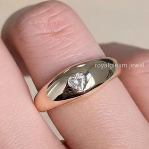 Puede incluir: Un anillo de oro con un diamante en forma de corazón engastado en el centro. El anillo está en un dedo, con un fondo borroso que presenta una flor blanca y un pequeño objeto dorado.