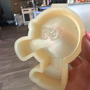 May include: A light yellow, Angry Birds-themed mold, featuring the character's face with prominent eyes and a beak. The mold is made of a translucent material and is designed for crafting or baking. The design is detailed and shows the character's features.