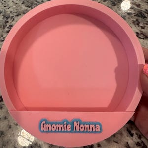 May include: A circular pink tray with a raised rim and a flat base. Attached to the front edge is a small, semi-circular pink tab featuring the text "Gnomie Nonna" in light pink letters set against a bright blue, rounded rectangular background.