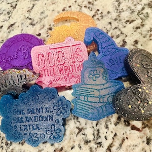 May include: A collection of colorful, glittery resin art pieces in various shapes. Includes a purple piece with text, a pink piece with a cross, a blue piece with a flower design, and a heart-shaped piece with text.