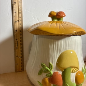 May include: A ceramic mushroom-shaped jar with a lid. The jar is off-white with vertical ridges. The lid is yellow-orange, and the jar is decorated with yellow, orange, and red mushrooms and green leaves. The jar is approximately 8 inches tall.