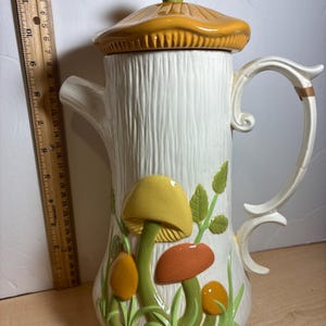 May include: A white ceramic coffee pot with a textured tree bark design. The pot features a lid and handle, and is decorated with colorful mushrooms and green foliage. The pot measures approximately 10 inches tall.