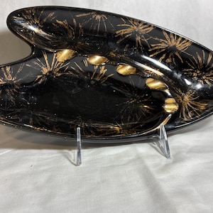 May include: A black ashtray with a unique, asymmetrical shape. The ashtray features a gold starburst pattern and gold cigarette rests. The glossy finish adds to its vintage appeal. A stylish piece for a retro home decor.