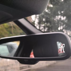Puede incluir: Retrovisor de coche negro con una calcomanía de lazo blanco. El espejo refleja un coche y una escena exterior borrosa. El lazo es un diseño simple y elegante.