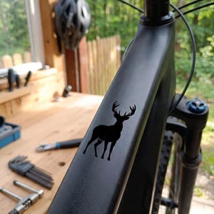 May include: A black mountain bike frame with a black deer silhouette decal. The bike has a matte finish and is set against a blurred background of a workshop and greenery. The handlebars and front suspension are visible.