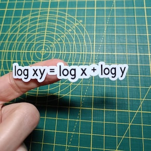 Puede incluir: Una pegatina blanca con la fórmula matemática "log xy = log x + log y" en letras negras. La pegatina está sujeta por un dedo sobre una esterilla de corte verde.