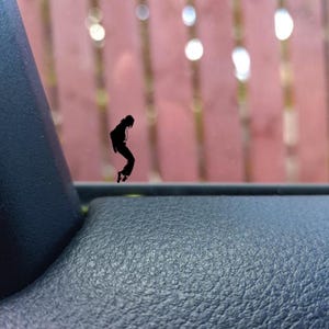 Puede incluir: Una silueta negra de una persona en pose de baile, sobre un fondo borroso de una valla roja y un interior de coche. La figura está en el aire, sugiriendo movimiento. La imagen es una pegatina o calcomanía.