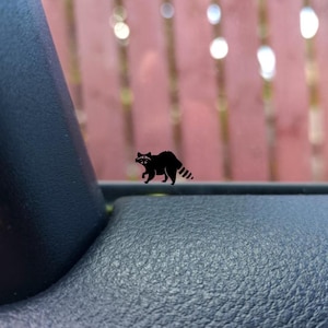 Puede incluir: Una calcomanía negra de mapache, con una silueta detallada del animal, está adherida a una superficie. El mapache se representa caminando, con su distintiva cola rayada visible. El diseño es simple, pero llamativo.