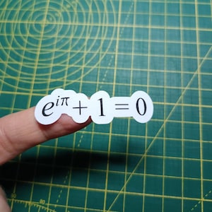 May include: A white sticker with the mathematical formula e^(iπ) + 1 = 0, held by a finger. The sticker has a rounded shape and black text. The background is a green cutting mat with grid lines.