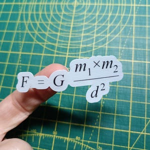 Könnte beinhalten: Ein hellblauer Aufkleber mit der Formel F = G (m1 x m2) / d2. Der Aufkleber wird zwischen den Fingern auf einer grünen Schneidematte mit Rasterlinien gehalten.