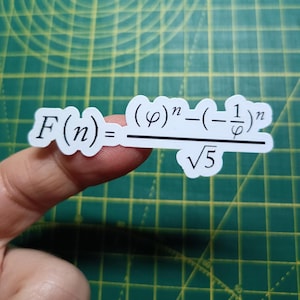 May include: A white sticker featuring a mathematical formula, held by a finger. The formula includes symbols, exponents, and a square root. The background is a green cutting mat.