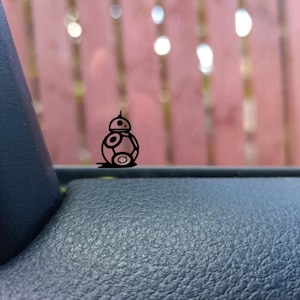 May include: A black vinyl decal of the Star Wars droid BB-8, featuring a round body and a small antenna. The decal is placed on a dark blue textured surface, with a blurred background of a wooden fence.