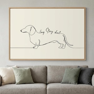 May include: A framed art print featuring a minimalist line drawing of a dachshund dog. The artwork is in black against a cream background, with the words "long story short" in script. The frame is a light wood color.