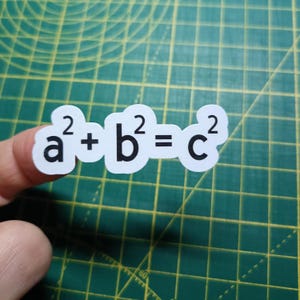 Può includere: Un adesivo bianco con la formula del teorema di Pitagora a² + b² = c² in lettere nere. L'adesivo è tenuto da un dito su uno sfondo a griglia verde.