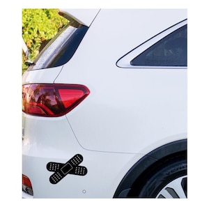 May include: A white car with a black bandage sticker on the rear. The sticker features two overlapping bandages with dotted patterns, a humorous and eye-catching design.