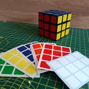 May include: A colorful Rubik's Cube with red, yellow, blue, and white squares. Several sets of replacement stickers in various colors are fanned out in front of the cube, ready for application. The cube sits on a green cutting mat.
