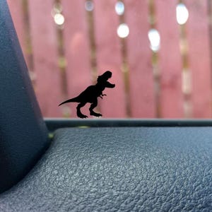 May include: Black Tyrannosaurus Rex dinosaur silhouette sticker. The sticker is on a window, with a blurred red background and bright, circular lights. The dinosaur is depicted in a running stance.
