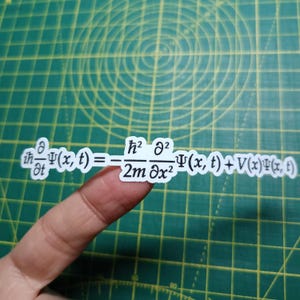 Puede incluir: Una pegatina blanca con una ecuación matemática en negro. La ecuación contiene símbolos y variables matemáticas. La pegatina está sujeta por un dedo sobre un fondo de cuadrícula verde.