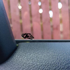 Può includere: Un adesivo nero del Millennium Falcon è applicato sul finestrino di un'auto. L'adesivo è una silhouette dettagliata dell'iconica astronave del franchise di Star Wars, su uno sfondo sfocato.
