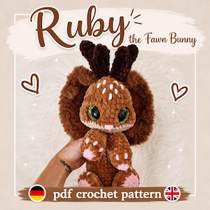 Könnte beinhalten: Ein braunes gehäkeltes Plüschhasenspielzeug namens Ruby the Fawn Bunny mit großen grünen Glitzeraugen, langen Ohren und kleinen Geweihstangen. Das Spielzeug hat weiße Punkte auf dem Kopf und rosa Akzente an den Pfoten. Text: Ruby the Fawn Bunny.