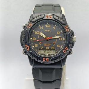 May include: Black Alba digital watch with a round face and a black rubber strap. The watch face features orange and white numerals, hands, and markers. The watch displays the time as 12:00 AL. The watch is water resistant to 10 BAR.