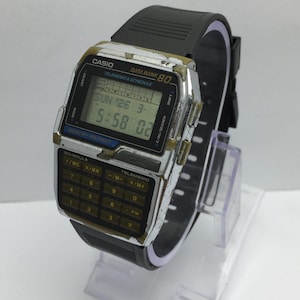 May include: A vintage Casio Data Bank 80 digital watch with a black rubber strap. The watch has a silver-colored face with a calculator keypad and a digital display showing the time and date. The watch is displayed on a clear plastic stand.