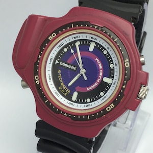 May include: A maroon-colored watch with a black band. The watch face features a white, black, and purple design with the text "GOOBERZ HI-TECH MASTER U.S.A." and "MICRO FLASH BEAM". The watch has a round face with a digital display.